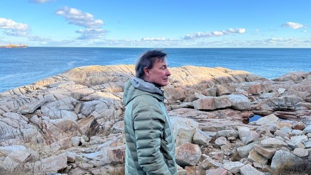 A man in a puffer jacket stands on rocks near the ocean.