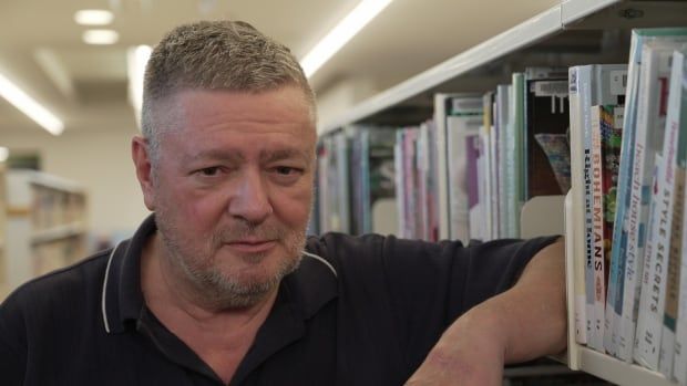 A middle aged man with grey stubble, wearing a blue polo shirt, leans on a bookshelf in a library