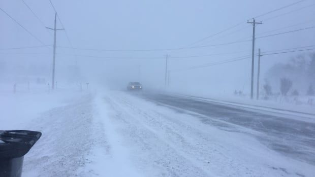 A road with snow-covered grass on either side and blowing snow, with a truck in the distance. 