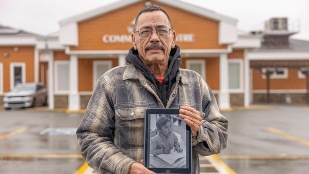 A man holds a black-and-white image of his young self, outside a community centre.