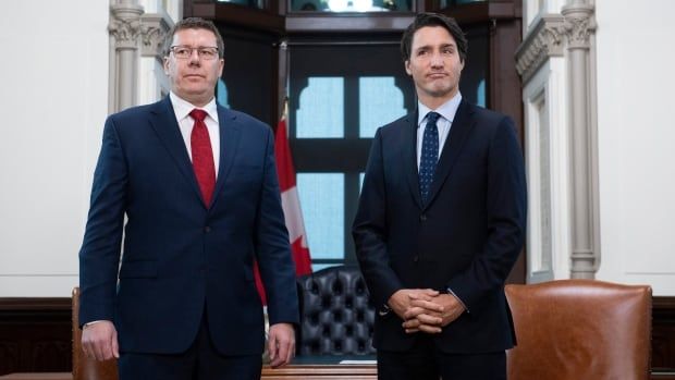 Two men in dark blue suits stand next to one another looking onward.