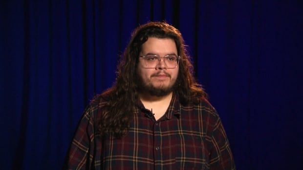 A white man with long brown hair and glasses in a red plaid shirt stands in a TV studio with blue lighting. 