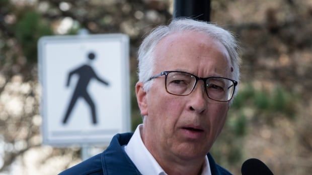 A man speaks into a mic outdoors, with a pedestrian walking sign behind him.