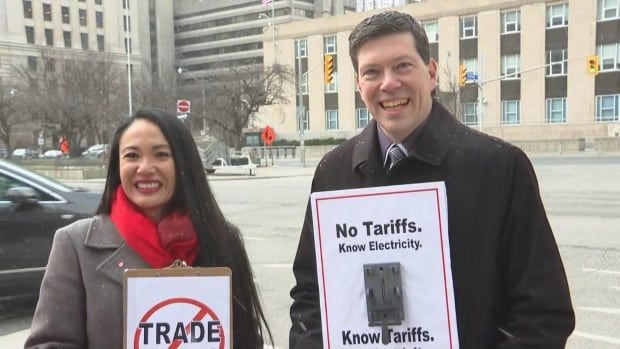 A man and a woman standing side-by-side holding anti tariff signs