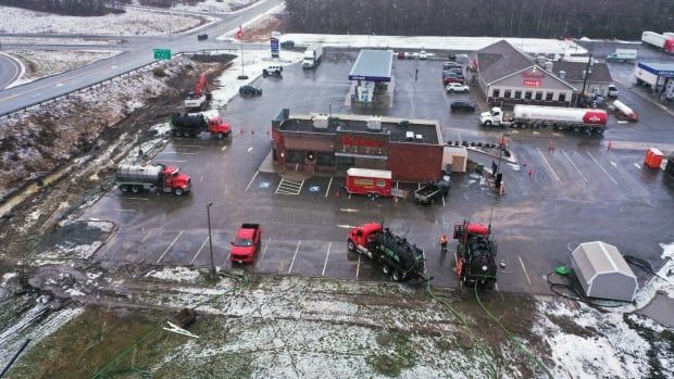 An aerial view of a Tim Hortons with four pump trucks surrounding it with green hoses running into the ditches nearby. Across the parking lot is in Irving oil gas station.