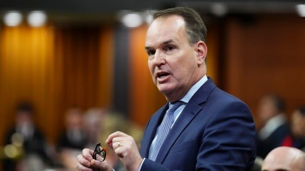 A minister stands in the House of Commons, wearing a navy suit and grasping a pair of reading glasses. 