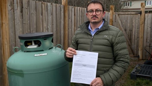 A man with glasses and a moustache stands next to a green propane tank. He is holding a piece of white paper.