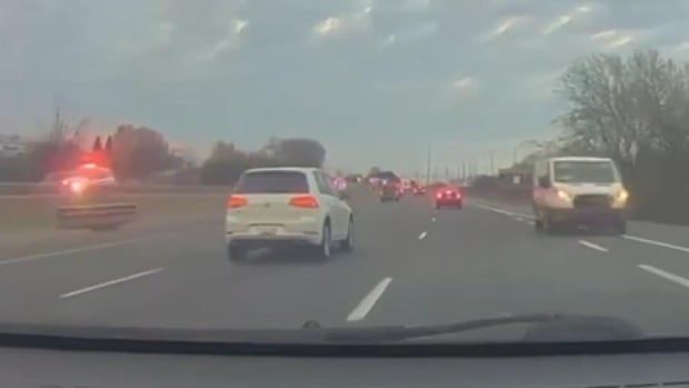 A white van on the shoulder of a highway. A police vehicle with lights flashing is on the other side of the median.