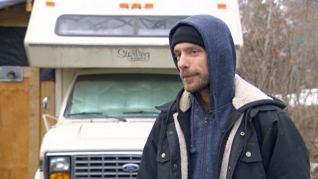 A white man with salt and pepper beard in a black toque, with a blue hooded sweatshirt pulled over his head and black jacket, stands in front of a white RV with bare trees in the background