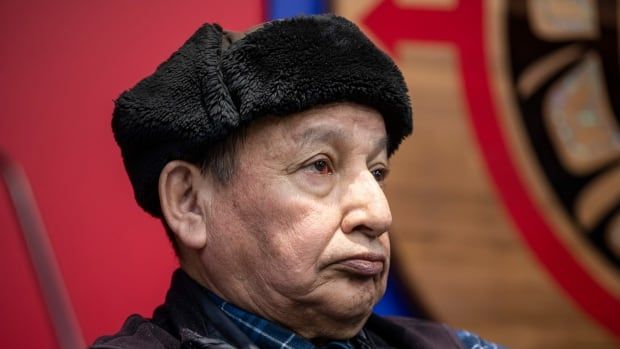 An Indigenous man wearing a black hat is pictured in profile, with a crest behind him.