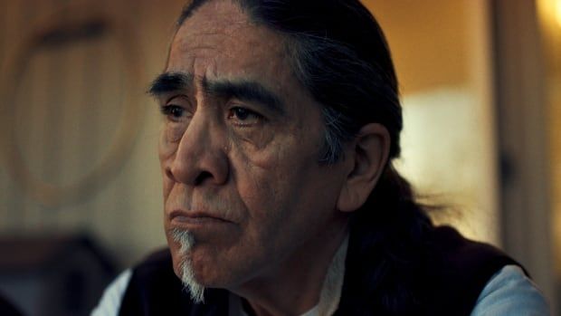 An older Indigenous man with busy eyebrows, a lined face and a thin white goatee looks into the distance.