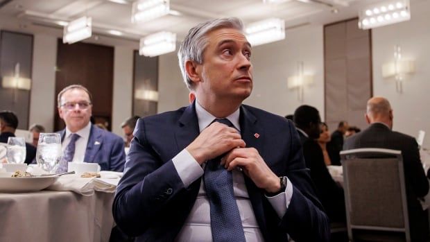 A man sits in a ballroom, adjusting his tie and looking to the side.