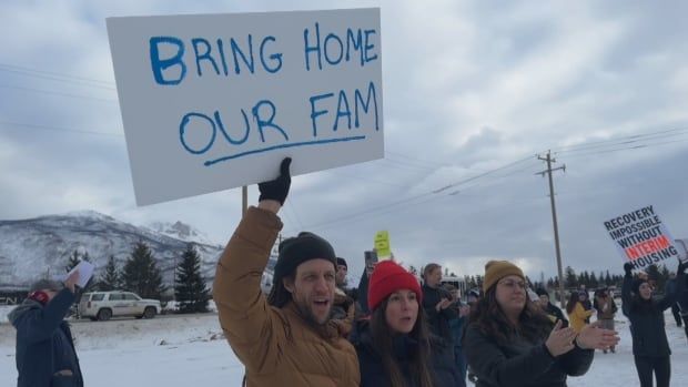 A man in a brown winter coat holds up a sign that reads, 'Bring home our fam.' Beside him are two women. Behind them is a larger crowd of protestors. They are all standing outside on snow-covered land, with mountains behind them.