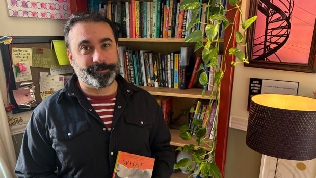 A man with a salt and pepper beard and dark hair in a black button-up shirt with the top three buttons undone revealing a red and white striped t-shirt underneath, holding a book in his home. A bookshelf is behind him. 