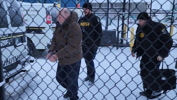 A man is seen through a fence walking in handcuffs with two sheriffs behind him 