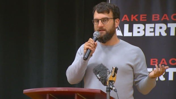 A man wearing a grey long-sleeved shirt speaks into a microphone in front of a podium.