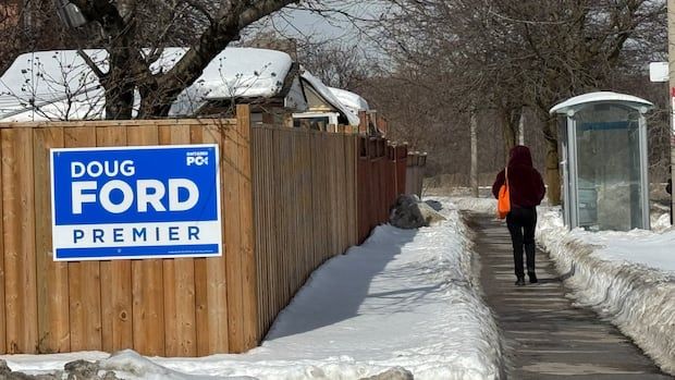 A Doug Ford campaign sign on a fence, left, while someone walks on a sidewalk nearby.