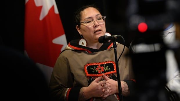 A woman stands at a microphone in a dark room, with a Canadian flag visible in the background.