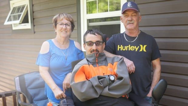 A woman in blue, a man in orange, grey and black in a wheelchair and a man in black stand looking at the camera on their porch. 