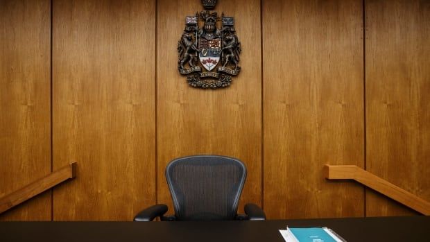 An empty chair where a judge usually sits on the bench in front of a wood-paneled wall bearing the provincial crest.