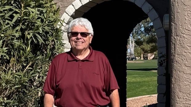 A man with white hair and black sunglasses stands outside in front of an arched doorway. A green tree and green grass are in the background.