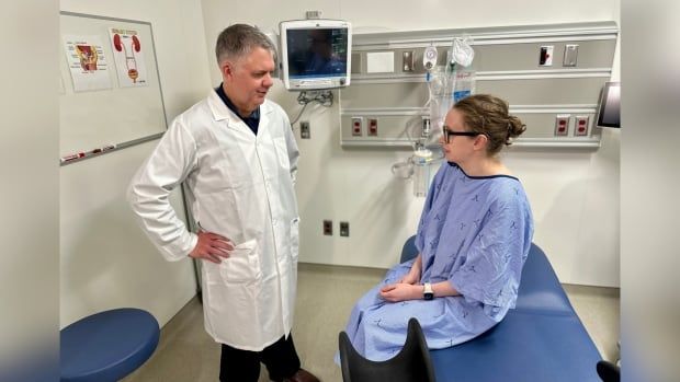 A doctor in a lab coat speaks to a patient dressed in a blue gown in a examination room.