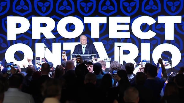 A man in a blue suit standing in front of a crowd. Behind him, is a screen that reads: "Protect Ontario."