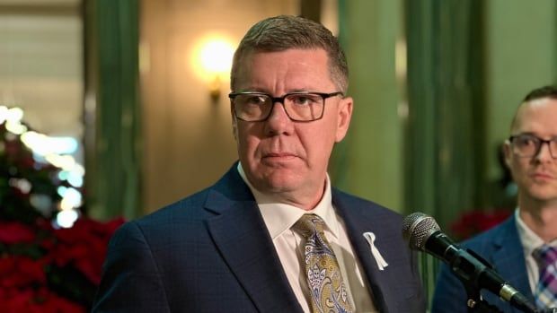 A man is wearing a white shirt, blue suit and gold and blue paisley tie. The man is standing in front of a microphone.