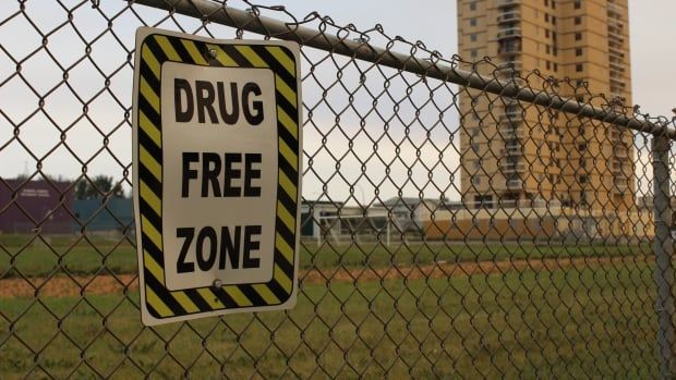 A sign on a fence. A school and a highrise can be seen in the distance.