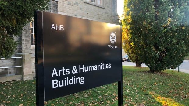 A black sign reads "arts and humanities building". In the background the side of a stone building is visible, and sunlight shines on green trees and grass on a unviersity campus