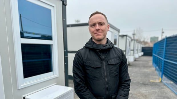 A portrait of a person in a windbreaker standing outside by a row of tiny cabins. 