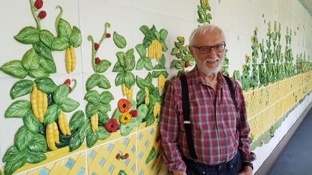 A man wearing a red plaid shirt and black suspenders stands in front of a tiled wall. The wall has green and yellow tiles on it and ceramic vegetables.