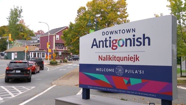 A white sign reading 'downtown Antigonish' and the Mi'kmaw name Nalikitquniejk underneath it, with cars driving by into the downtown core and colourful buildings along the street