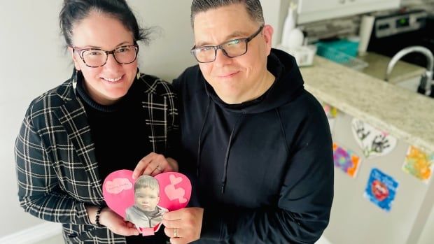 A man and a woman stand side by side holding a paper heart with their son's picture in the middle.