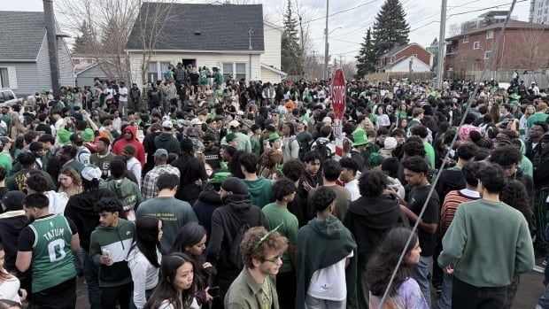 A crowd of green-clad partygoers stand around houses and stop signs on a street. 