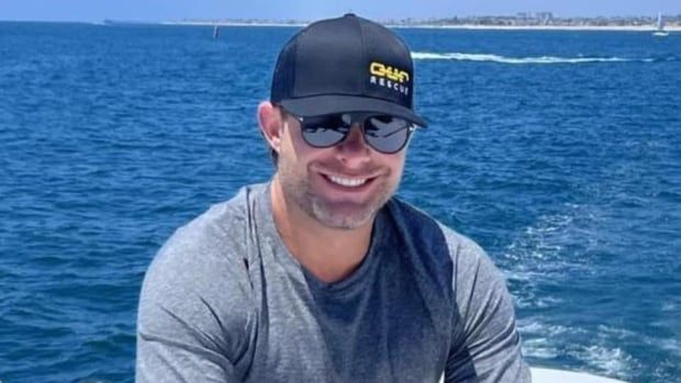 A man wearing sunglasses and a ball cap in swim trunks and a sweater sits in a boat on a white bench, smoking a cigar and smiling broadly at the camera while the vast expanse of a deep blue sea is laid out behind him.