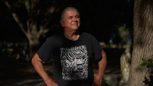 A man with gray hair and a black graphic T-shirt stands outside next to a tree, his hands on his hips. 