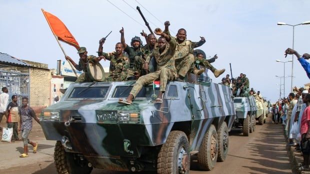 Several men in military fatigues raise their arms and salute atop a tank being driven down a road in what appears to be a parade. Onlookers watch or cheer on either side of the road.