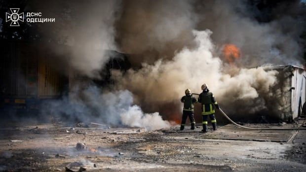 Two firefighters in full uniform and helmet are shown with their backs to the camera utilizing a firehose as massive clouds of smoke and some orange fire are shown.