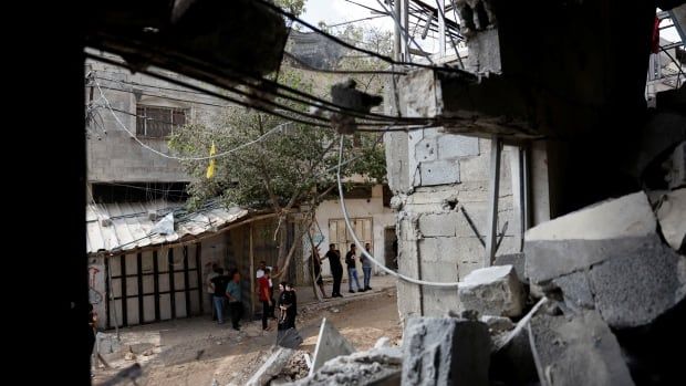 Several people stand on a ruined street, photographed through an opening in a building, with rubble surrounding the hole in the structure. 