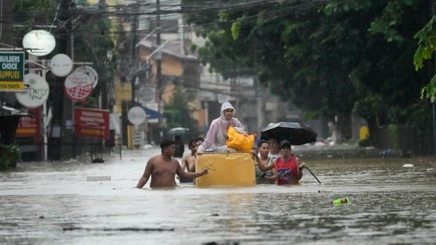 A group of people wade through chest-high water on a flooded street. One person sits on a floating yellow object in the centre of the group. 