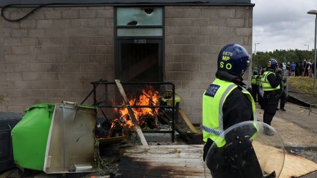 Two helmeted and outfitted police officers are shown standing near a low-rise building, where a small fire is shown.