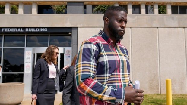 A large, bearded, dark-complected man in a collared striped shirt holds a water bottle while walking outside a building. A woman in a suit jacket and skirt is shown in the background.