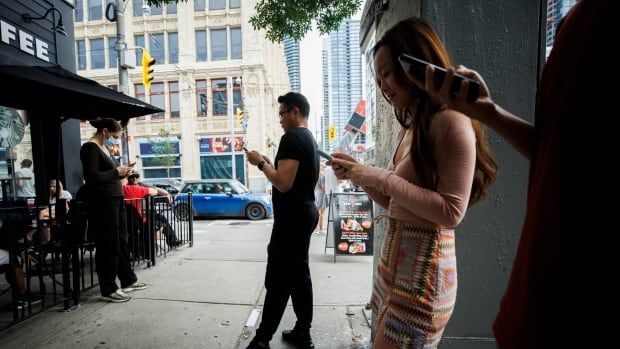 Men and women stand outside a coffee shop on a busy city street. They are all looking at their phones. 