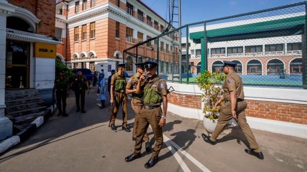 Half a dozen police officers in green uniforms stand together on the street on a sunny day.