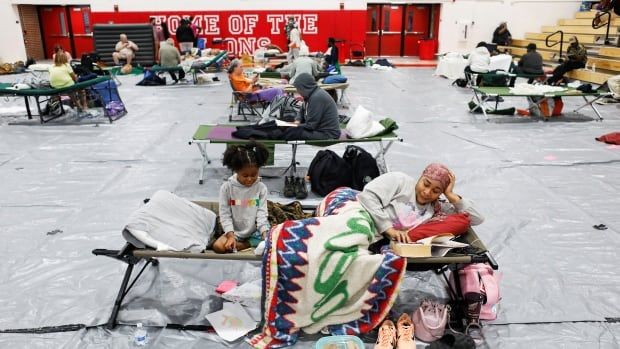 People sit and lie on cots in a gymnasium.