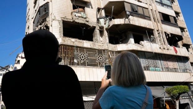 The backs of two people are shown in the foreground staring up at a multistorey building in a city that is heavily damaged, with windows blown out.