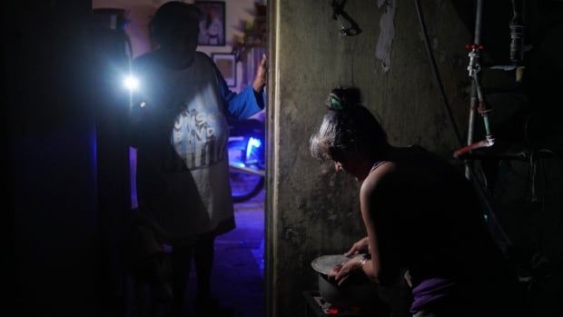 A person cooks on a stove amid a blackout as another person standing in the doorway uses a mobile phone's flashlight to illuminate the kitchen.