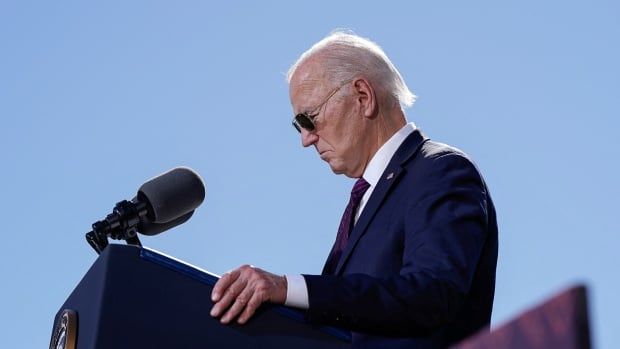 A man in a suit and aviator sunglasses looks down solemnly at a podium.