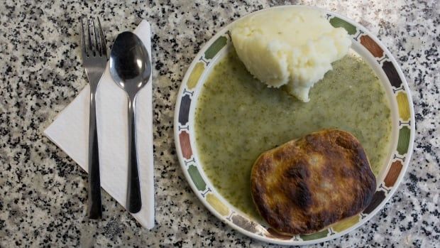 a plate of mashed potatoes and a meat pie sit, smothered in a green sauce, on a grey speckled counter beside a fork and knife